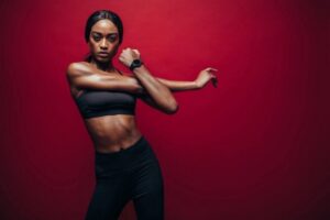 Athletic woman in a black sports bra and leggings stretching with arms crossed against a red background.
